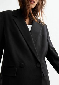 Blazer nero doppiopetto con rever a scialle, due tasche anteriori e bottoni rotondi. Tessuto liscio con design su misura.