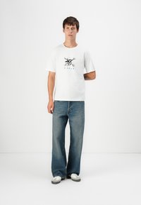 Hvid t-shirt med et kraniumgrafik med hat og korslagte knogler, parret med løse blå denimjeans og grå sneakers.
