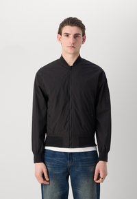 Schwarze Bomberjacke mit Reißverschluss, gerippten Bündchen, Seitentaschen und glatter Textur. Trügt über einem weißen T-Shirt und blauen Jeans.