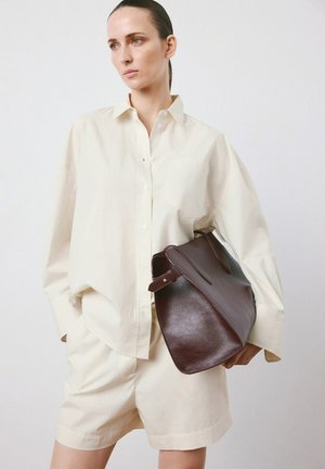 Woman in oversized cream shirt and matching shorts holding large dark brown leather handbag against white background.