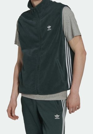 Man draagt een donkergroene Adidas mouwloze vest met rits over een grijs T-shirt en bijpassende Adidas broek met witte strepen.