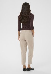 Pantalon beige sur mesure à jambe fuselée, présentant une texture lisse et une taille élastique. Porté avec un haut foncé ajusté et des mocassins noirs.