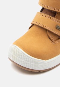 Pantofii din suede bej cu două curele cu arici, talpă din cauciuc alb, detaliu texturat pe capacul degetelor și accente de cusătură.