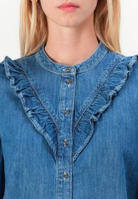 Denim shirt in blue with a round collar, button front, and ruffle accents along the placket and shoulder seams. Soft texture.