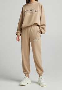Felpa beige e pantaloni coordinati in tessuto morbido. La felpa presenta un testo stampato; i pantaloni hanno una stampa simile vicino alla tasca.