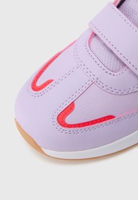 Fioletowy sneaker z czerwonymi akcentami i teksturowaną tkaniną, wyposażony w pasek na rzep i zakrzywiony kształt. Podeszwa wykonana z brązowej gumy.