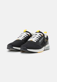 Baskets en mesh noires avec des lacets blancs, une boucle jaune au talon et une semelle grise avec un accent orange.