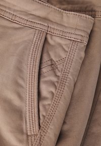 Hnědá bavlněná tkanina s prošívanými švy a malou kapsou. Textura je hladká a design zahrnuje vyztužené okraje.