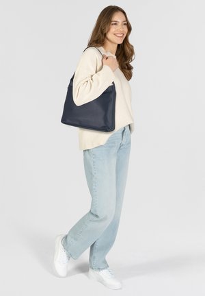 Bolso tote de cuero azul marino con una textura suave, que presenta un diseño minimalista y una forma blanda y relajada. Modelo vistiendo un suéter ligero y vaqueros.