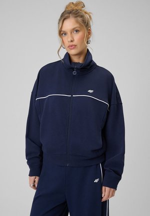 Młoda kobieta z blond włosami związanymi w kok, ubrana w granatową sportową kurtkę i spodnie 4F z białymi lamówkami na jasnoszarym tle.