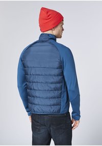 Blaue isolierte Jacke mit gesteppter Rückseite und glattem Stoff vorne, kombiniert mit einer roten Mütze. Verfügt über lange Ärmel und einen hohen Kragen.