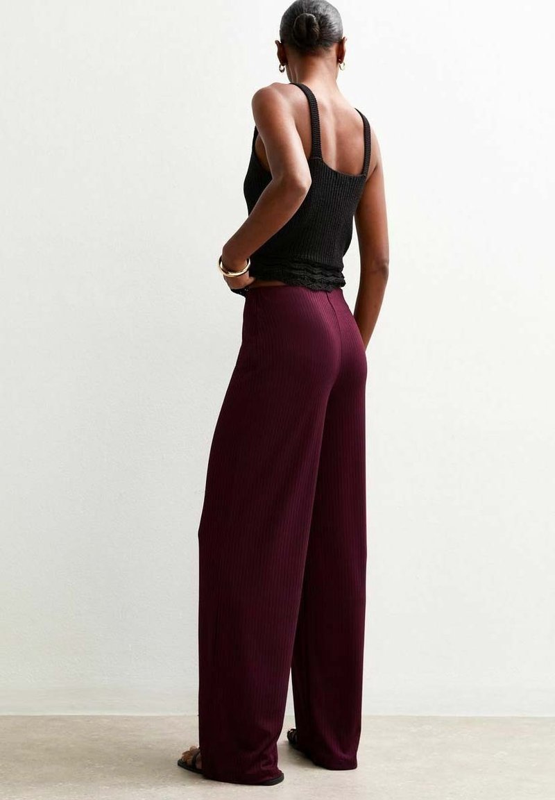 Femme aux cheveux en chignon, portant un haut noir sans manches et un pantalon bordeaux ample à taille haute, debout contre un mur uni.