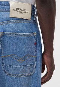 Πίσω όψη ενός ατόμου που φοράει ανοιχτογάλανα τζιν Replay με ραμμένη τσέπη και λευκή δερμάτινη ετικέτα της μάρκας στη μέση.