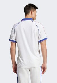Biely športový polokošeľ s modrými a červenými akcentmi na golieri a okrajoch rukávov; priliehavý strih, krátke rukávy, hladká textúra.