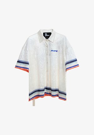 Hombre con rastas que lleva pantalones cortos blancos y camisa grande a juego con rayas azules y naranjas, diseño de ojo azul en la espalda y calcetines blancos.