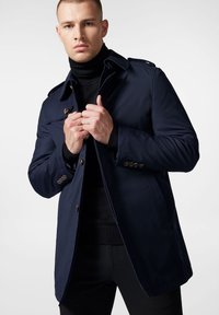 Granatowy trencz z podwójnym zapięciem, pagonami na ramionach i guzikami. Wykonany z gładkiego, lekkiego materiału.