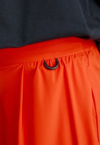 Oransje plisserte skjørt med en glatt tekstur, som har en svart løkke detalj nær midjen. Bakgrunnen er mørk.