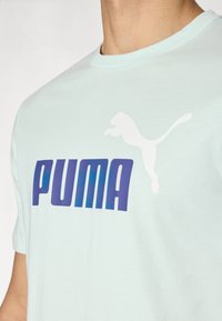 Šviesiai mėlynas medvilninis marškinėlis su baltu Puma logotipu ir ryškiu purpuriniu užrašu. Minkšta tekstūra, apvalus iškirpimas ir trumpomis rankovėmis.