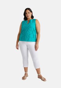 Top turchese senza maniche con un intricato motivo a taglio, abbinato a pantaloni bianchi a tre quarti e sandali color nocciola, che evidenziano una silhouette rilassata.