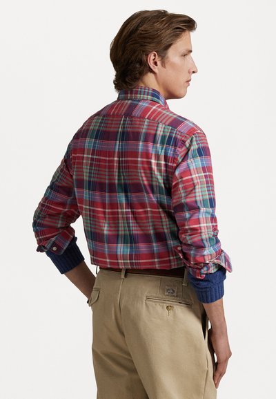 Mönstrad skjorta i flera färger med röd, blå och grön nyans, med blå ribbade ärmslut och button-up krage. Bärs tillsammans med beige byxor.