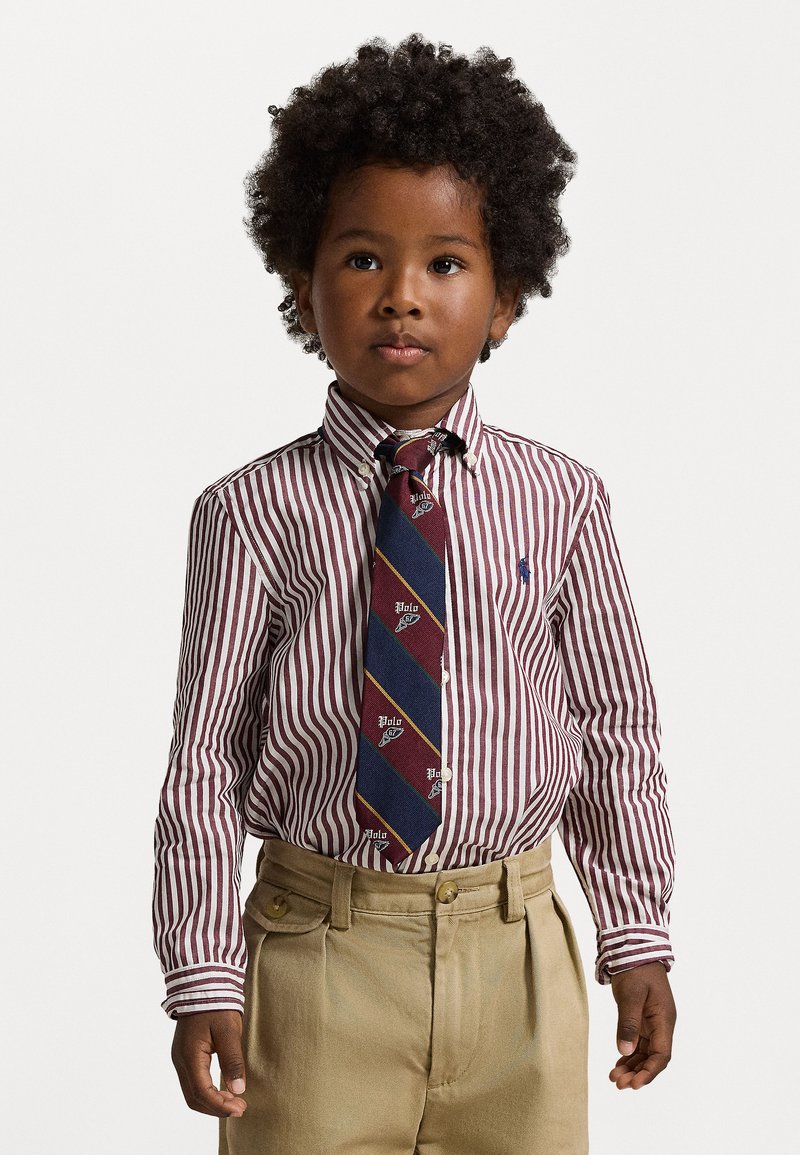 Nošama maroon un balto svītraino krekla ar tumši zilu kaklasaiti, kas rotāta ar dimanta rakstu, un khaki biksēm. Strukturēta auduma.