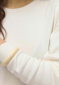 Kreminės spalvos, gumburuota megzta ilgos rankovės palaidinė su vientisu tekstūru. Ant riešo uždėtas šviesios spalvos bangle.