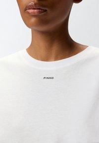 Bela bombažna majica s krožnim izrezom, na prsih z majhnim natisnjenim črnim logotipom "PINKO". Gladka tekstura, preprost dizajn.