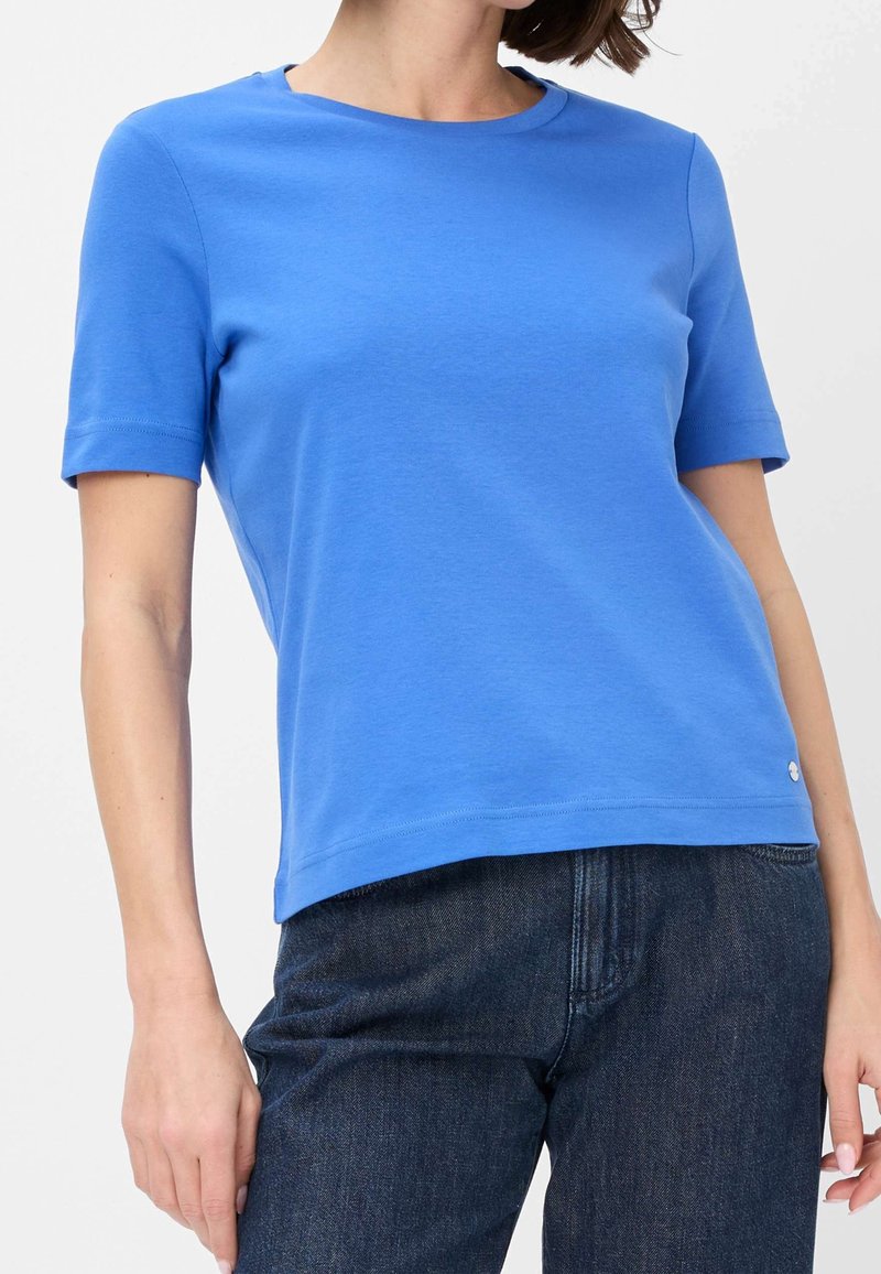 Femme portant un t-shirt bleu uni à manches courtes et col rond, légèrement rentré dans un jean bleu foncé sur fond blanc.