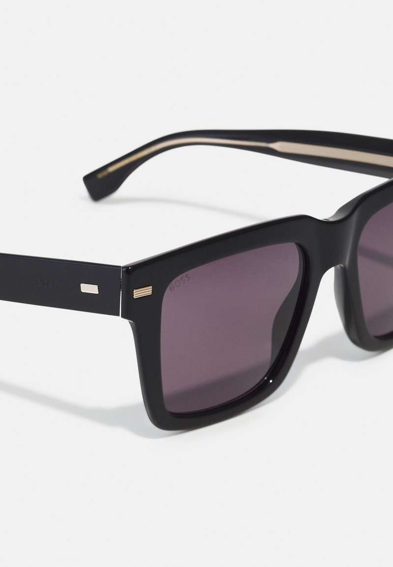 Lunettes de soleil noires surdimensionnées avec des verres carrés. La monture mate présente des accents dorés sur les branches et un logo discret près de la charnière.