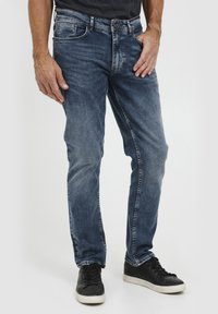 Jeans in denim blu scuro con vestibilità dritta, dotati di cinque tasche e una sottile texture sbiadita. Indossati con sneakers nere alte.
