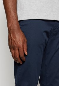 Pantaloni blu scuro realizzati in tessuto liscio, che mostrano cuciture dettagliate. Una mano poggia sulla coscia, mostrando vene e texture della pelle visibili.