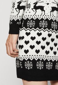 Pull noir et blanc présentant un motif de rennes, de cœurs et de flocons de neige, avec des poignets et un ourlet côtelés, affichant un design en tricot texturé.