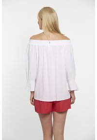 Weiße schulterfreie Bluse mit gerafftem Ausschnitt und langen Ärmeln, kombiniert mit roten Shorts. Glattes Material und geraffte Ärmelbündchen.
