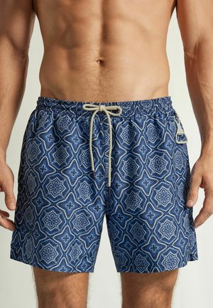 Torse masculin portant un short de bain bleu avec un motif géométrique floral blanc et un cordon noué à la taille.