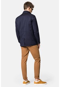 Granatowa wodoodporna kurtka z stójką, tylnym wentylowanym panelem i subtelnym detalem z logo, zestawiona z jasnobrązowymi spodniami i żółtymi trampkami.