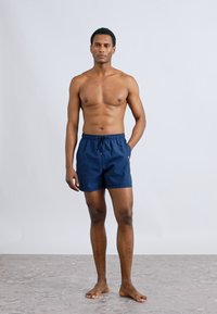 Μπλε σκούρο μαγιό σορτς από ελαφρύ ύφασμα, με ελαστική μέση και κορδόνι για ρύθμιση. Το μοντέλο στέκεται ξυπόλυτο μπροστά σε ανοιχτό φόντο.