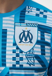 Tricou de fotbal albastru cu modele geometrice, guler rotund și un logo central al clubului cu textul "Droit au But" și o stea deasupra.