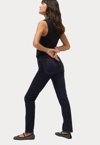 Jeans oscuros con costuras rojas contrastantes, diseño ajustado, pierna acampanada, combinados con unos zapatos planos negros con lunares y una blusa sin mangas.