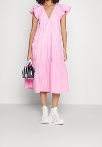 Różowa, warstwowa sukienka bawełniana z falbaniastymi rękawami i głębokim dekoltem w serek, w połączeniu z białymi, masywnymi sneakersami i małą czarną torebką.