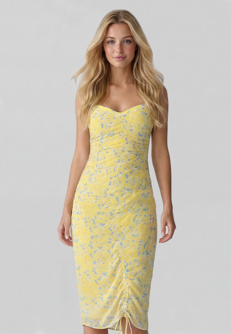 Femme aux longs cheveux blonds portant une robe jaune ajustée sans manches avec un motif abstrait bleu, debout devant un fond clair uni.
