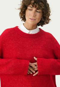 Röd stickad tröja med bred ribbad halsringning och långa ribbade muddar. Modellen har en vit skjorta med krage under. Slen textur.