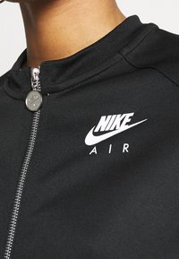Detailní záběr horní části hrudníku osoby, která má na sobě černou zipovou bundu Nike Air se stříbrným táhlem zipu a bílým logem na levé straně.