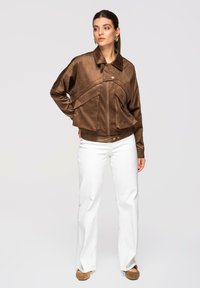 Femme portant une veste en cuir marron oversize avec poches à rabat, un pantalon blanc droit et des chaussures marron, debout devant un fond uni.