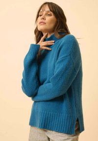 Turkusowy sweter z wysokim kołnierzem, wykonany z miękkiej, teksturowanej dzianiny. Posiada ściągacze przy mankietach i u dołu oraz boczne rozcięcia dla większej swobody.