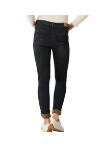 Femme portant un jean skinny bleu foncé retroussé et des baskets blanches, les mains posées sur les hanches, vue de dos.