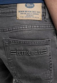 Pantaloni in denim grigio con un'etichetta testurizzata con il marchio "Petrol" e dettagli di cucitura, inclusa una tasca posteriore accentuata da testo in rilievo.