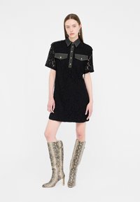 Kvinde iført en kortærmet sort blondekjole med knappede brystlommer og knæhøje støvler med slangehudsmønster, stående mod en hvid baggrund.