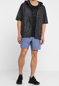 Svart kortärmad jacka med huva och zip-stängning, bärs över en svart t-shirt, i kombination med blå shorts och svarta sportskor.