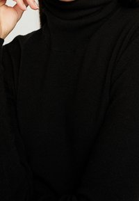 Pull à col roulé noir en maille douce, avec un col montant et des manches longues. Gros plan mettant en valeur la texture et la coupe.