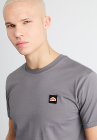 Graues Baumwoll-T-Shirt mit Rundhalsausschnitt und einem kleinen, gestickten Patch in Schwarz, Orange und Rot auf der linken Brustseite.
